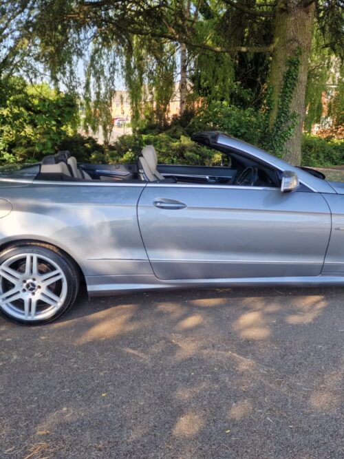 Side view of a 2012 Mercedes-Benz E-Class E250 AMG Cabriolet in metallic silver with the roof down, parked on a sunny day in the UK
