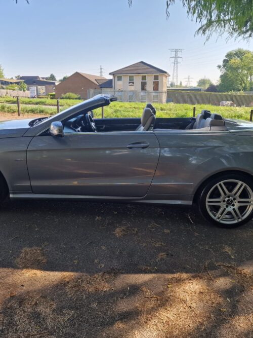 Full side profile of a 2012 Mercedes-Benz E220 Cabriolet in metallic silver with AMG alloy wheels and convertible top down, parked on tarmac with trees and buildings in the background