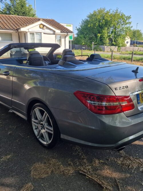 Rear-side view of the 2012 Mercedes-Benz E220 Cabriolet with the roof down, finished in metallic silver, showcasing the AMG alloy wheels and sleek tail lights