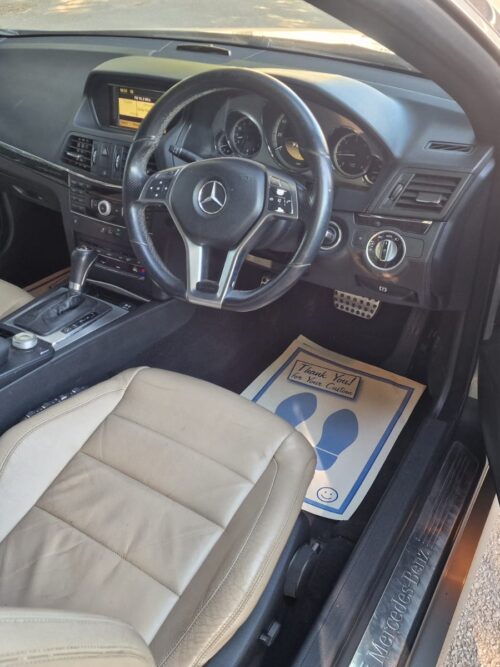 Driver-side view of the 2012 Mercedes-Benz E-Class E250 Cabriolet interior featuring beige leather seats, AMG styling, and multifunction steering wheel