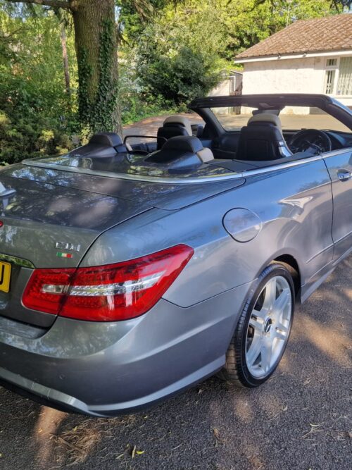 Rear-side view of a 2012 Mercedes-Benz E220 CDI Cabriolet in metallic silver with UK registration plate, showcasing the convertible's sleek design and twin exhausts