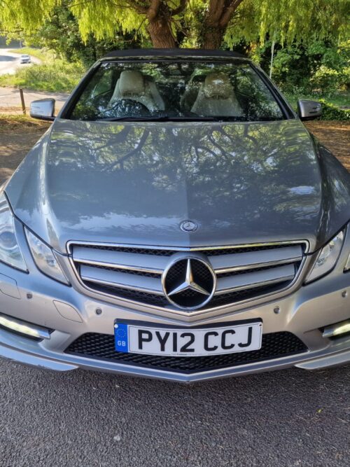 Front view of a silver 2012 Mercedes-Benz E-Class Cabriolet with roof down, parked under trees – for sale by Vaz Auto UK.